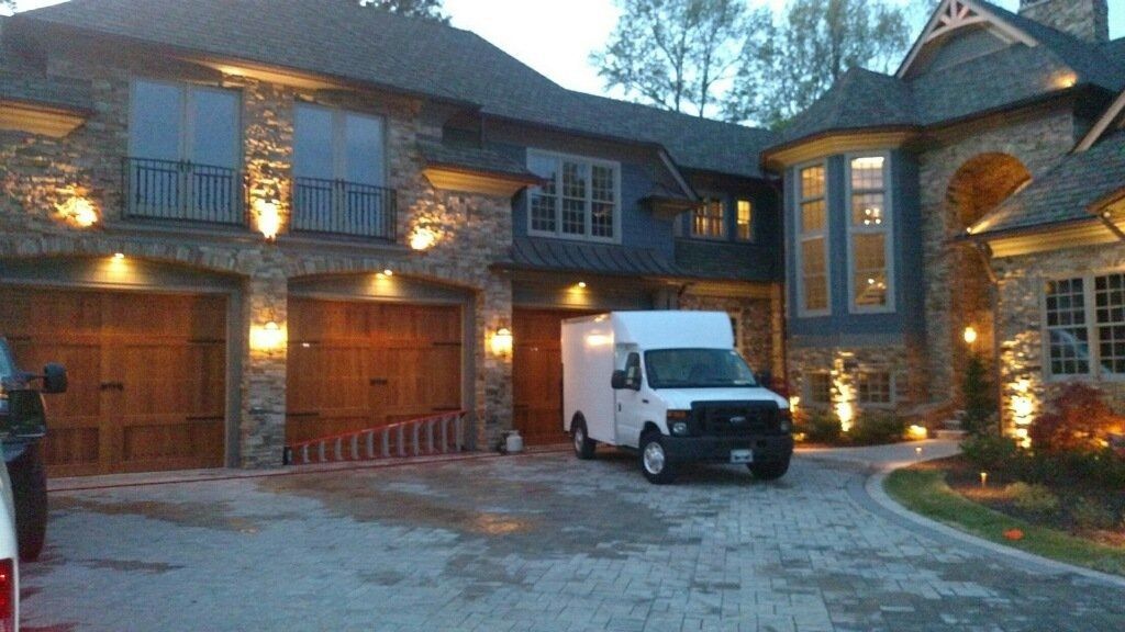 Large stone mansion at dusk with lit exterior lights, garage doors, and a white van parked in the driveway.