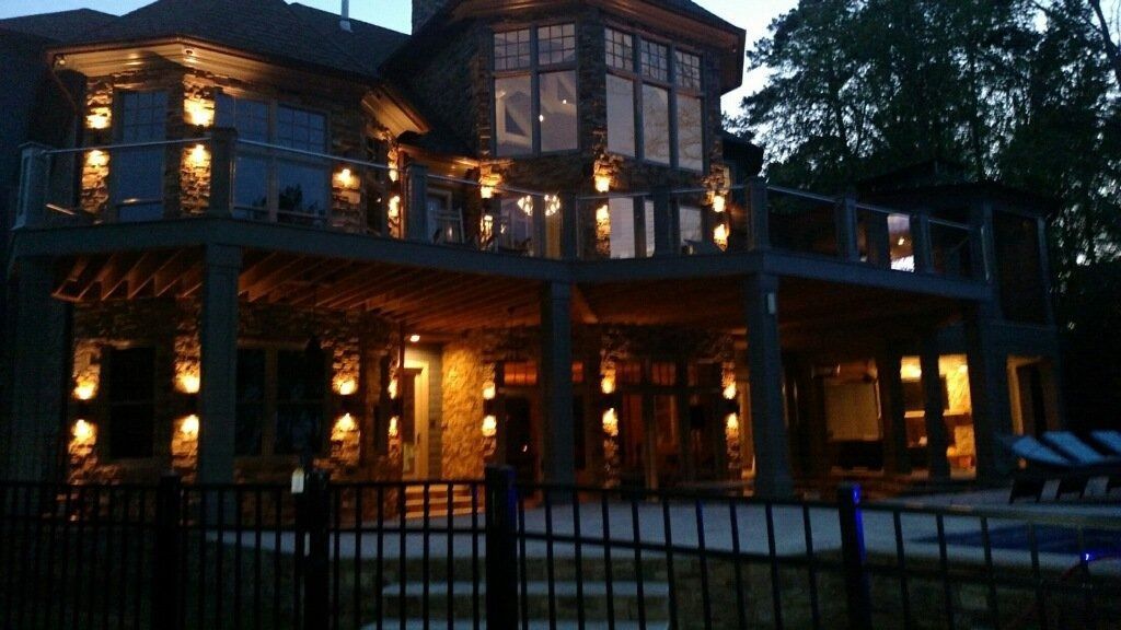 Lit multi-story house at dusk with warm interior lights and a fenced yard