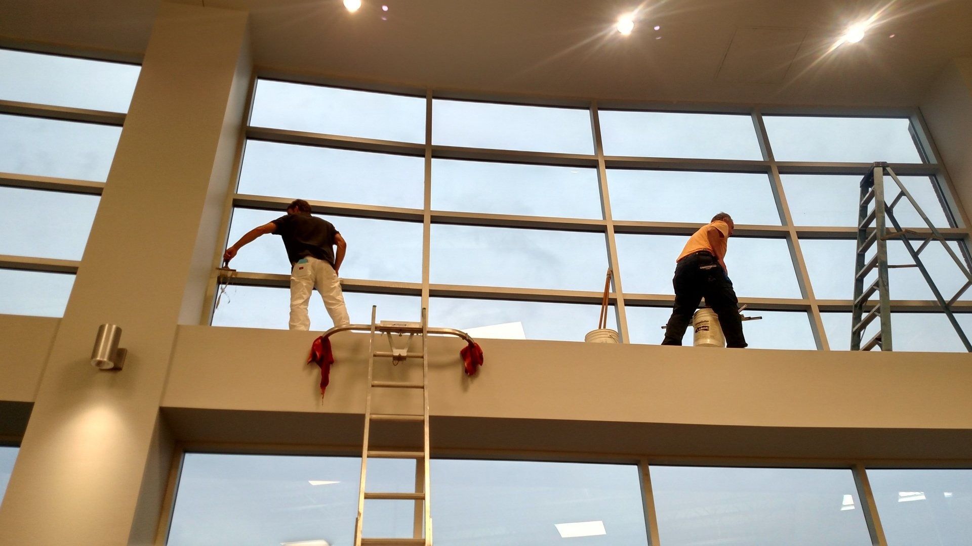 Workers cleaning a tall glass window wall from ladders inside a bright atrium