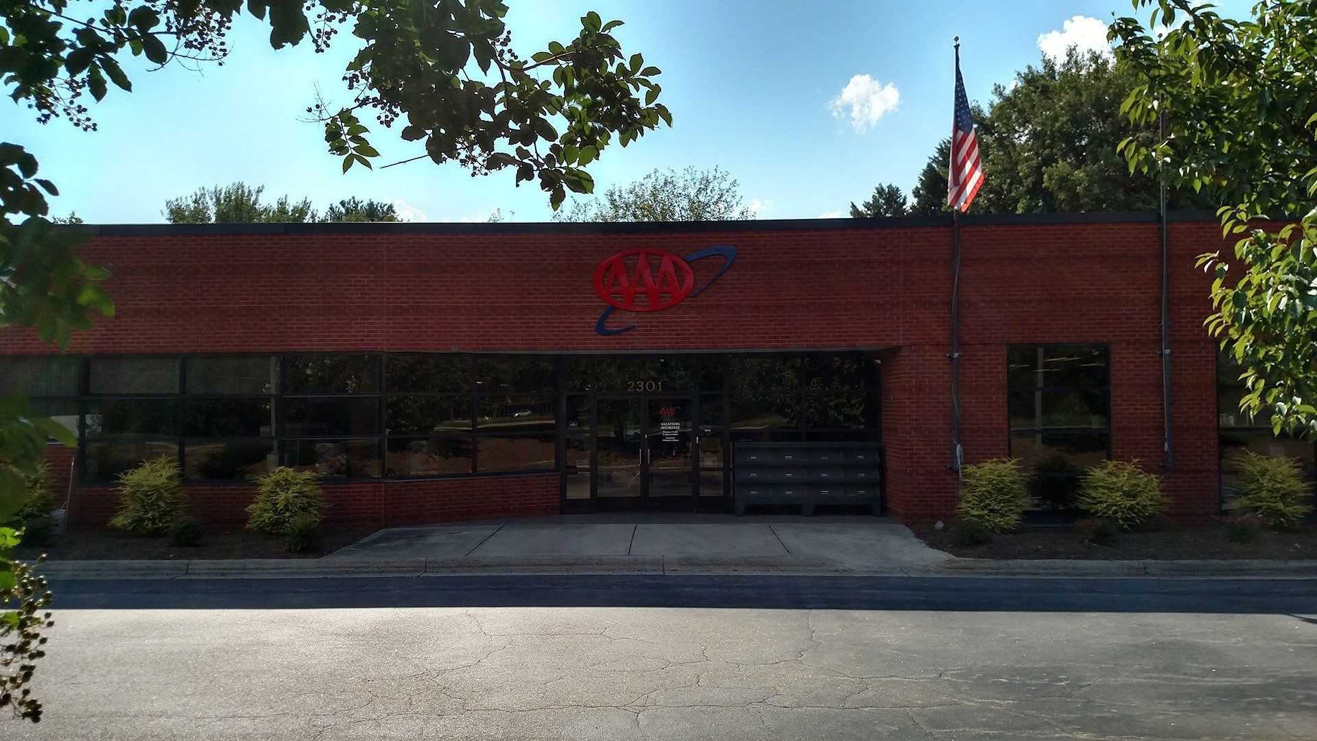 Brick building with a red sign above glass doors, shaded by trees beside a parking lot