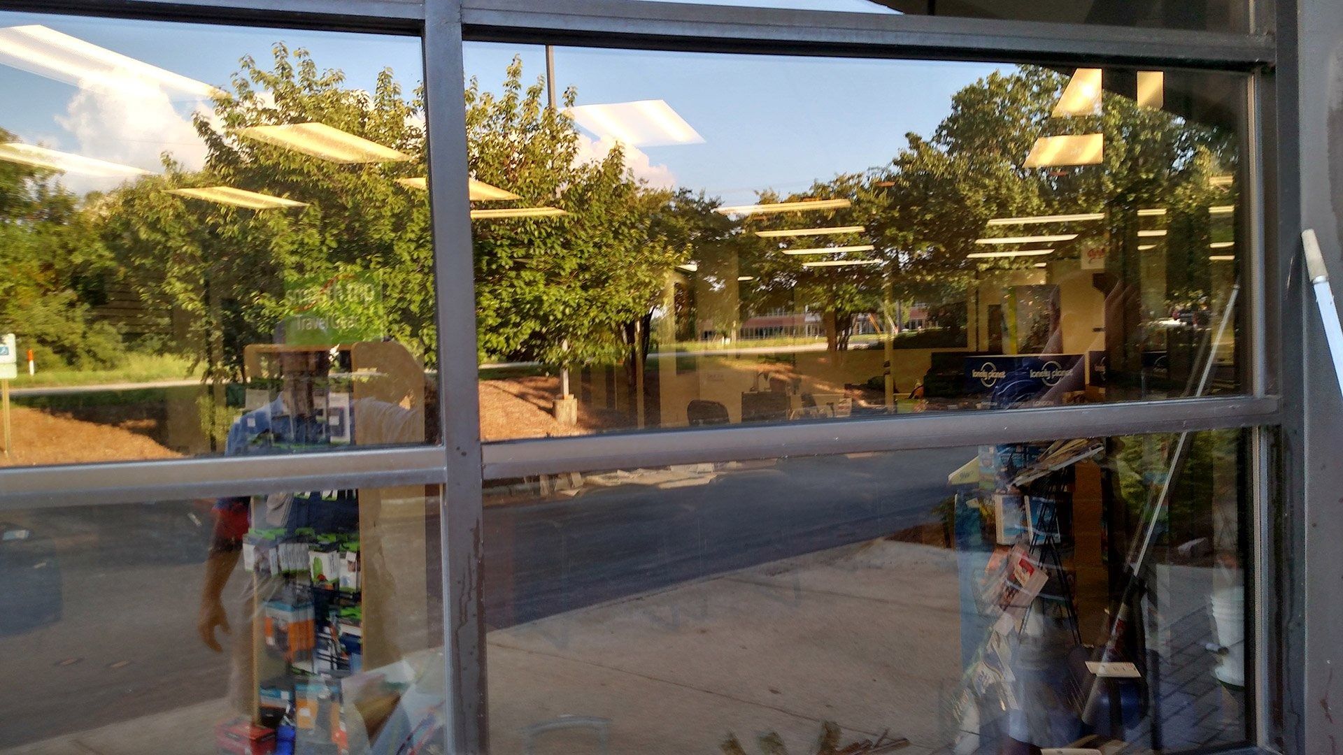 Storefront window reflecting trees and street outside, with shelves of colorful items inside.