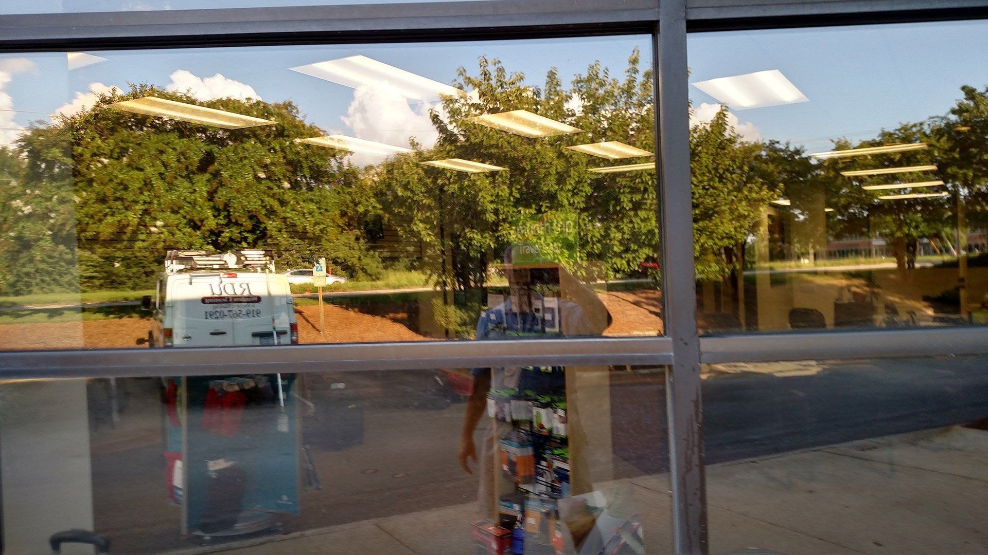 Storefront window reflecting trees and sky, with a sidewalk and shelves visible inside.