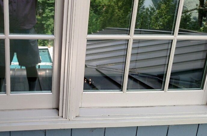 White-framed window reflecting trees, with a rolled-up gray shade inside and blue siding below