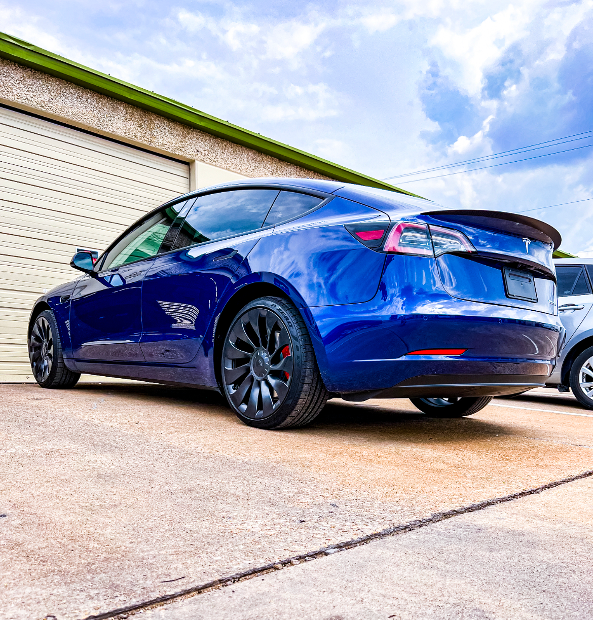 Blue Tesla sedan parked outside a building with a white garage door, on a sunny day.