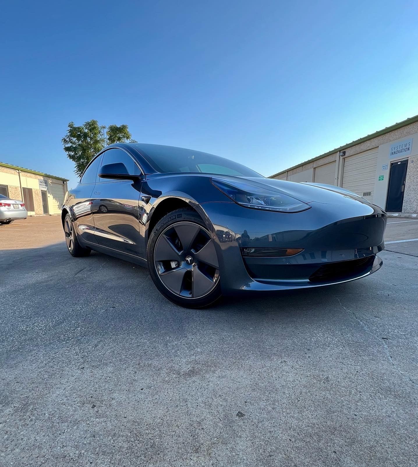 Dark blue Tesla car parked on asphalt, with buildings and blue sky in the background.