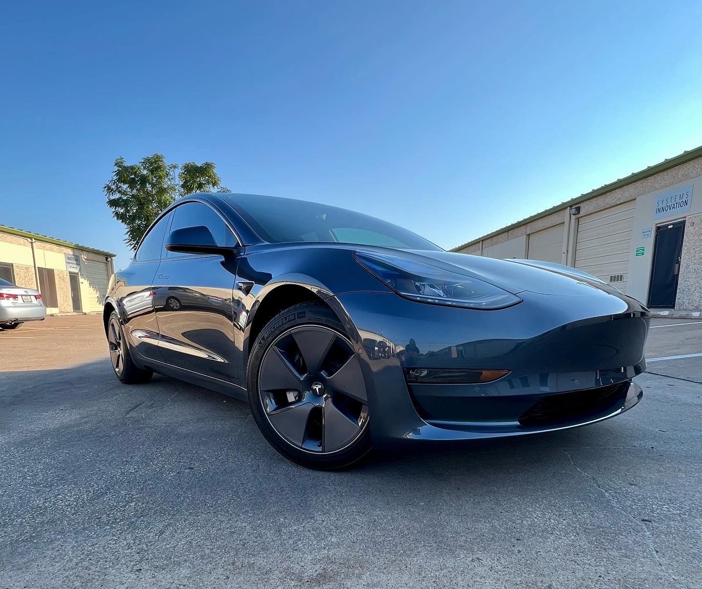 Blue Tesla Model 3 car parked outdoors under a bright blue sky.