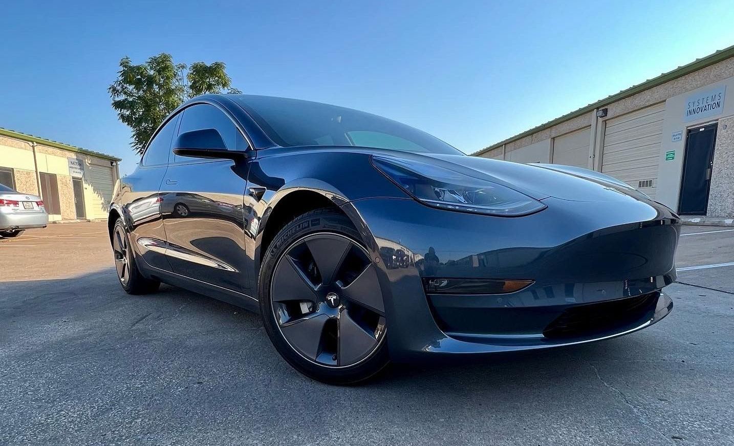 Blue Tesla Model 3 parked on asphalt in front of buildings on a sunny day.