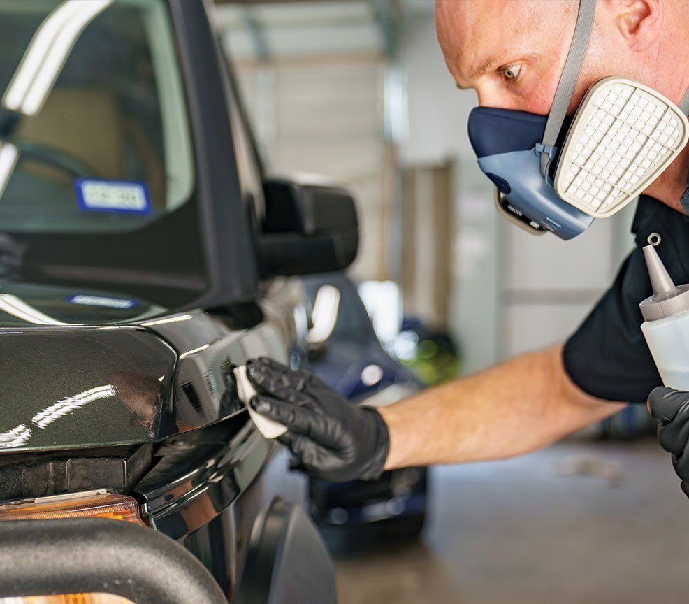 Person in respirator applying detailer to black car, wearing gloves.