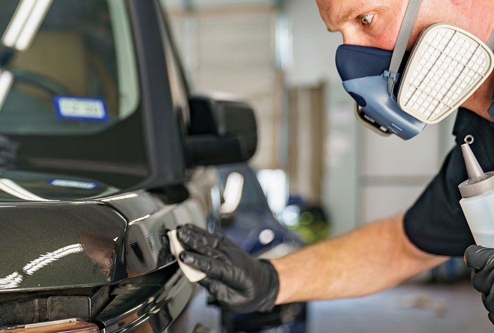 Man wearing respirator and gloves polishing black car with applicator in hand.