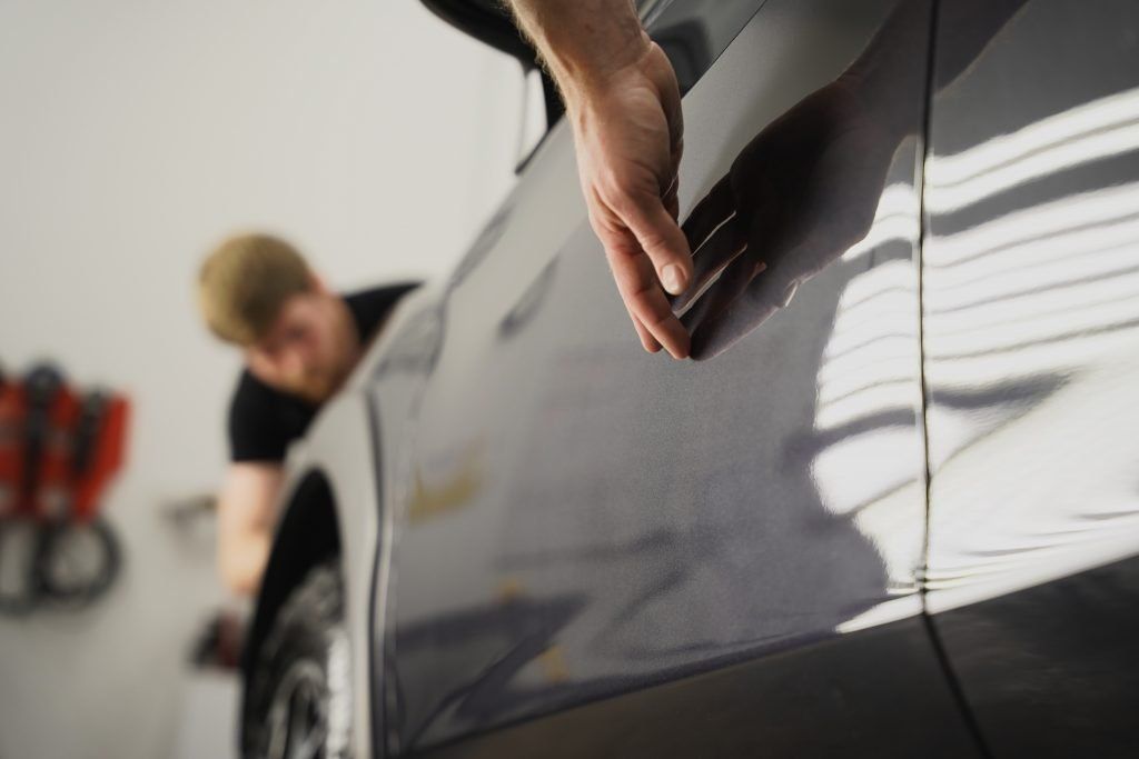 Person examining car bodywork in a garage, hand on the car's surface.