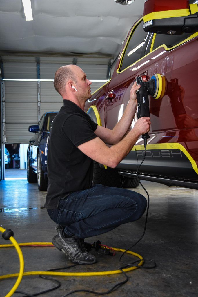 Man polishing a red car in a garage with a power tool, wearing dark clothing. Yellow tape protects areas.
