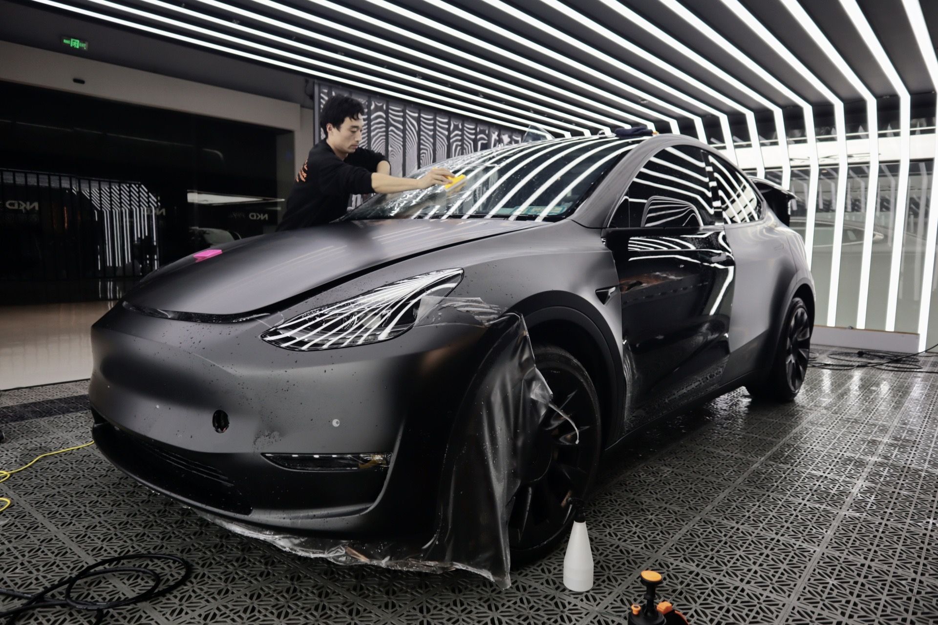 Man applying film to a matte black Tesla Model Y in a car detailing shop.