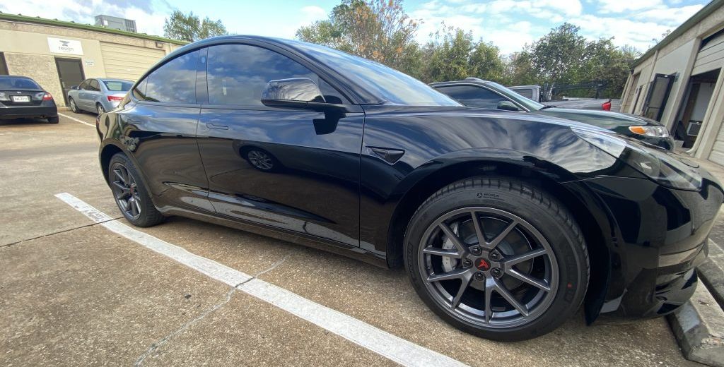 Black Tesla Model 3 parked, angled view; dark gray wheels, tinted windows, in a parking area.