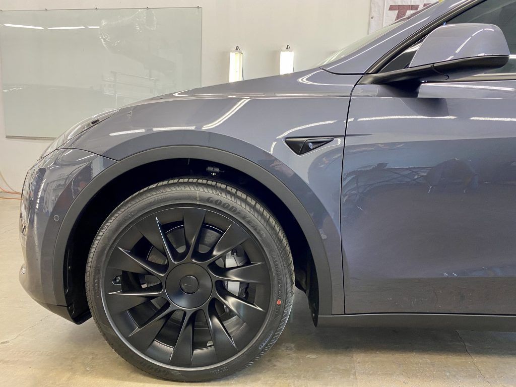 Gray Tesla Model Y car's front side, showing a black wheel and fender detail, in a brightly lit garage.