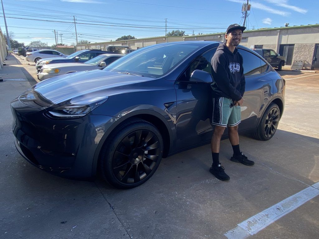 Man standing next to a dark blue Tesla Model Y; sunny outdoor setting.