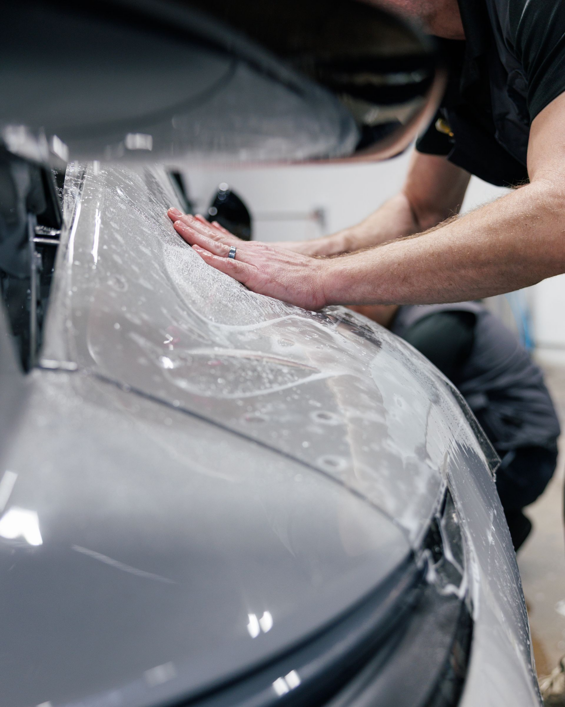 Person applying protective film to a car bumper; hands pressing down on the clear material.
