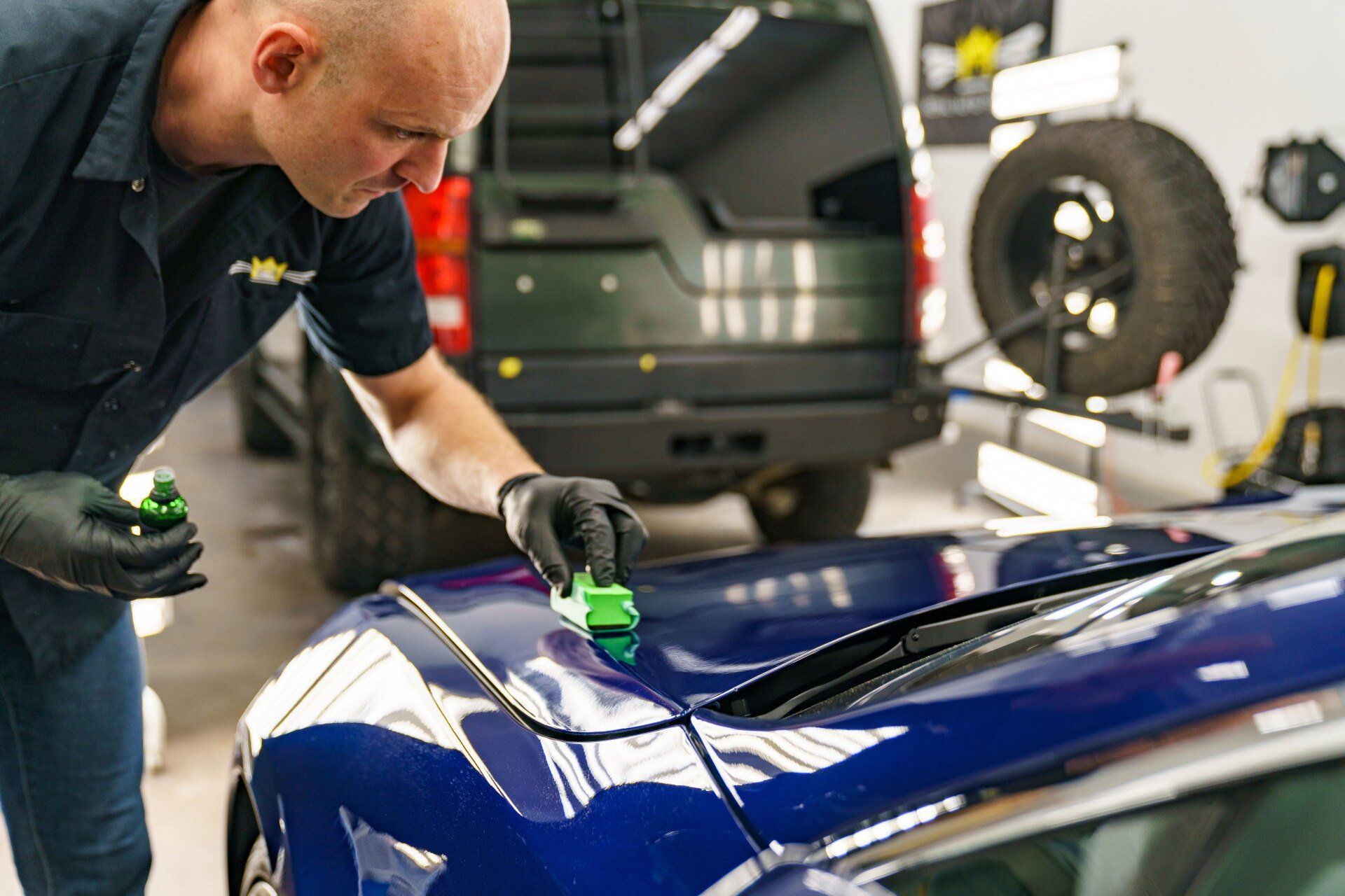 Man applying a green sealant to a blue car hood in a garage.
