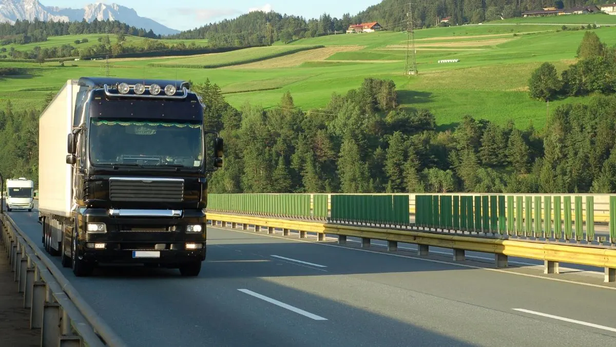 Black semi-truck driving on a bridge, green hills and trees in the background.