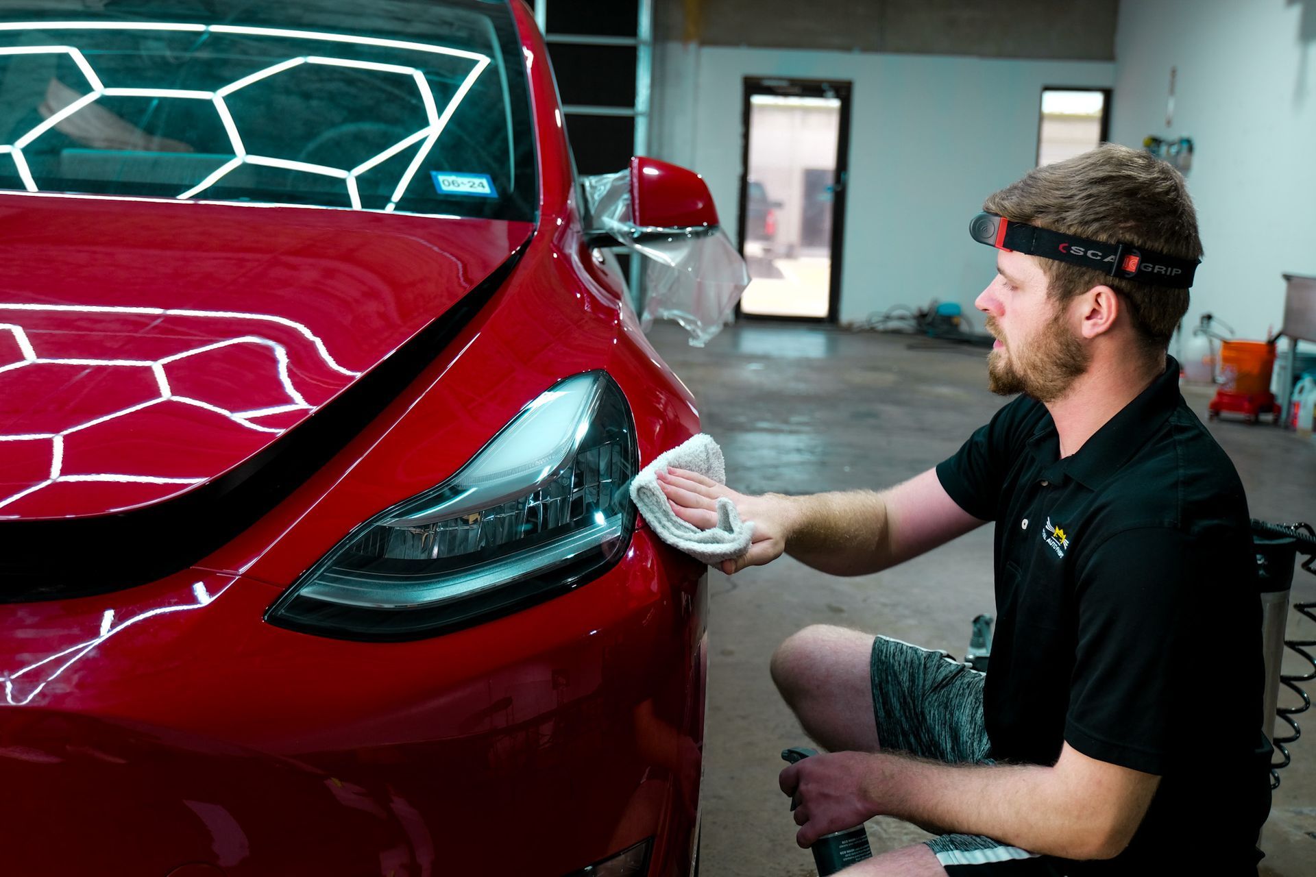 Man cleaning a red Tesla car's headlight inside a garage. He is wearing a headlamp.