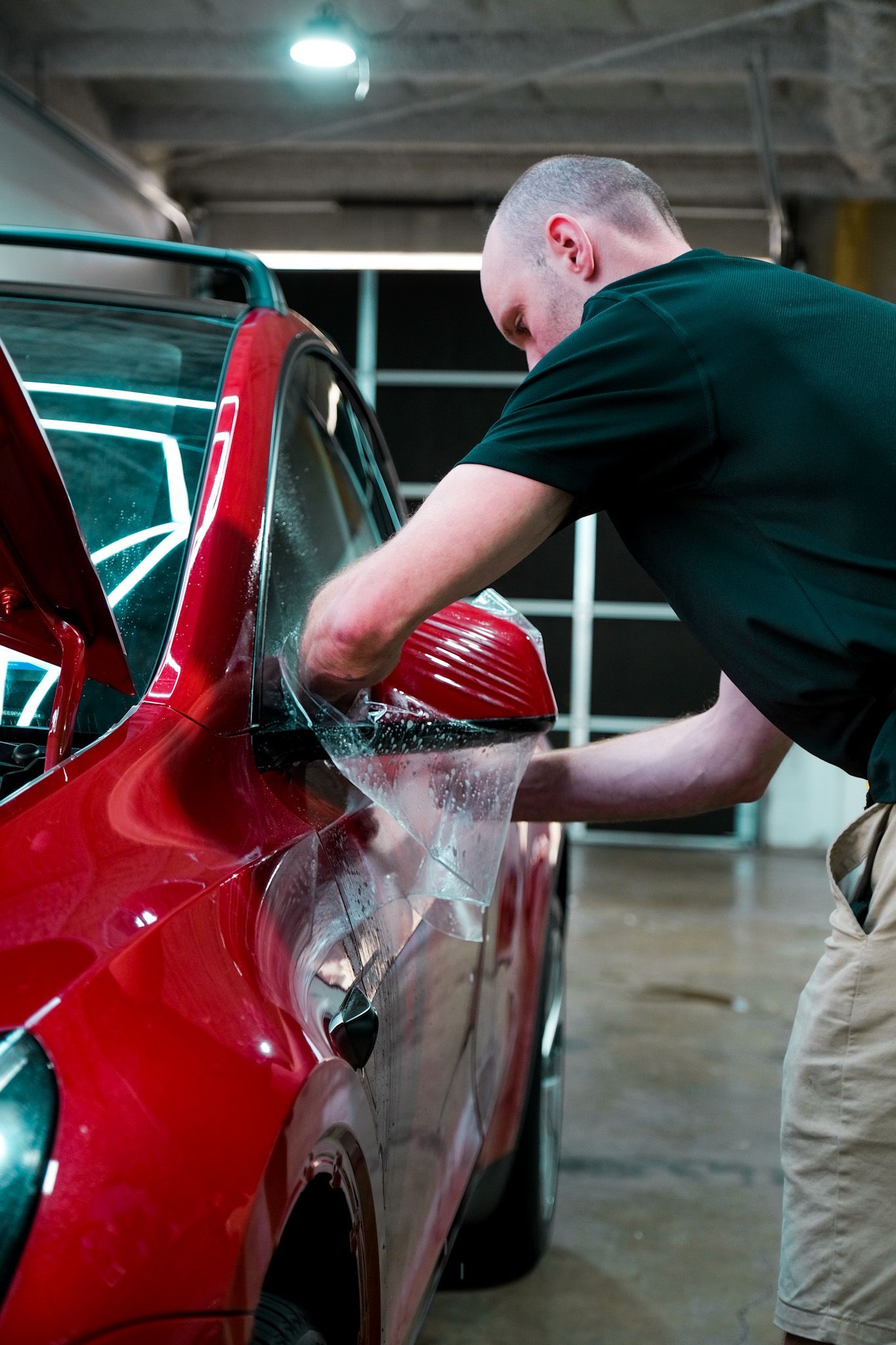 Man applying film to a red car's side mirror in a workshop.