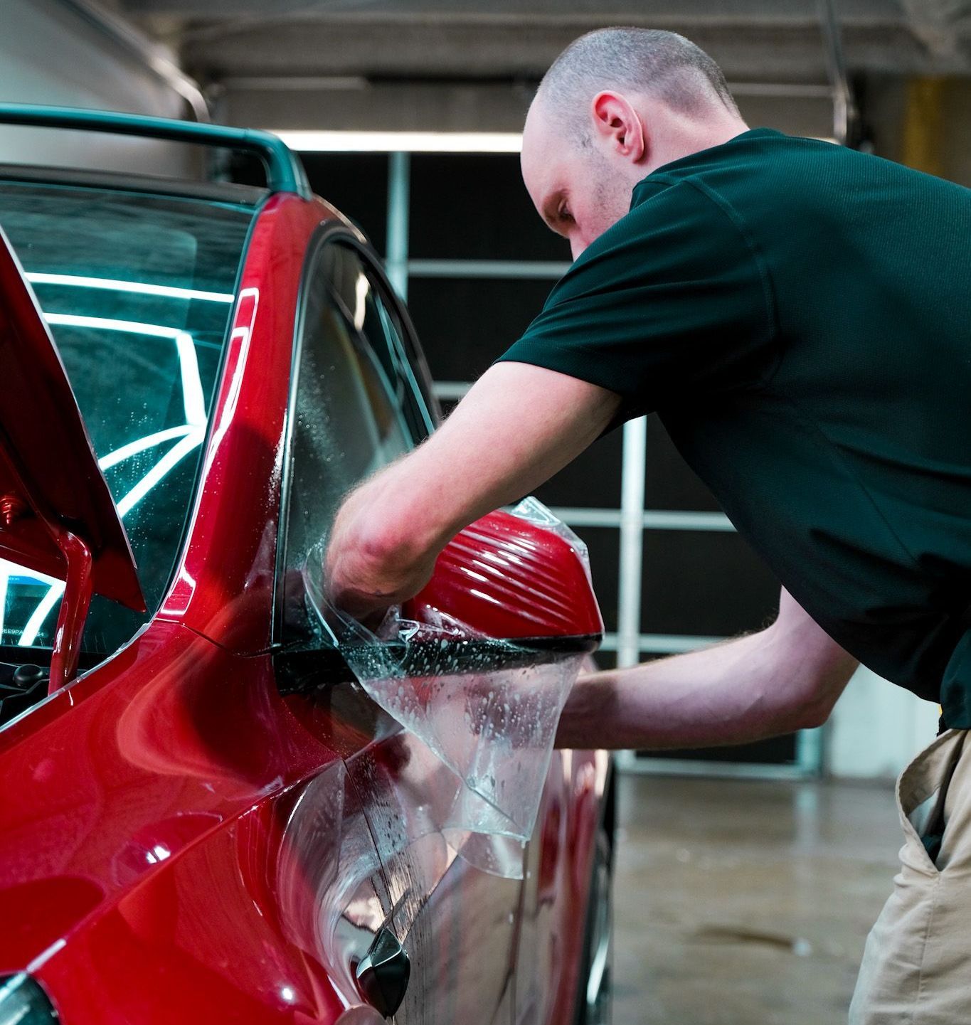 Man applying protective film to a red car's side mirror in a workshop.