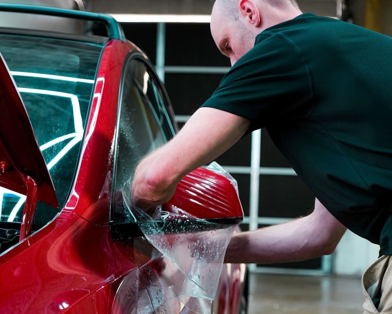 A person applying clear protective film to a red car's side mirror in a well-lit garage.
