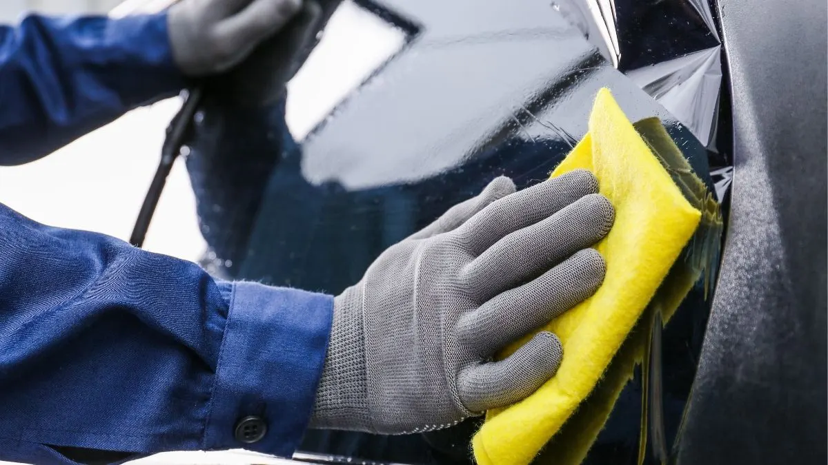 Person wearing gloves wiping a car's surface with a yellow cloth.