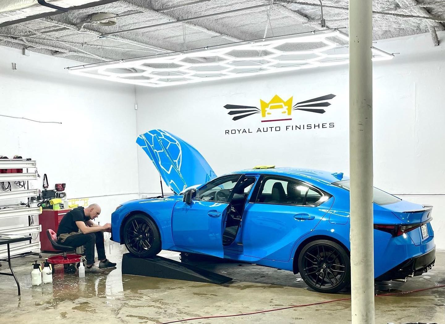 A mechanic works on a blue car in a bright garage. The hood and driver-side door are open.