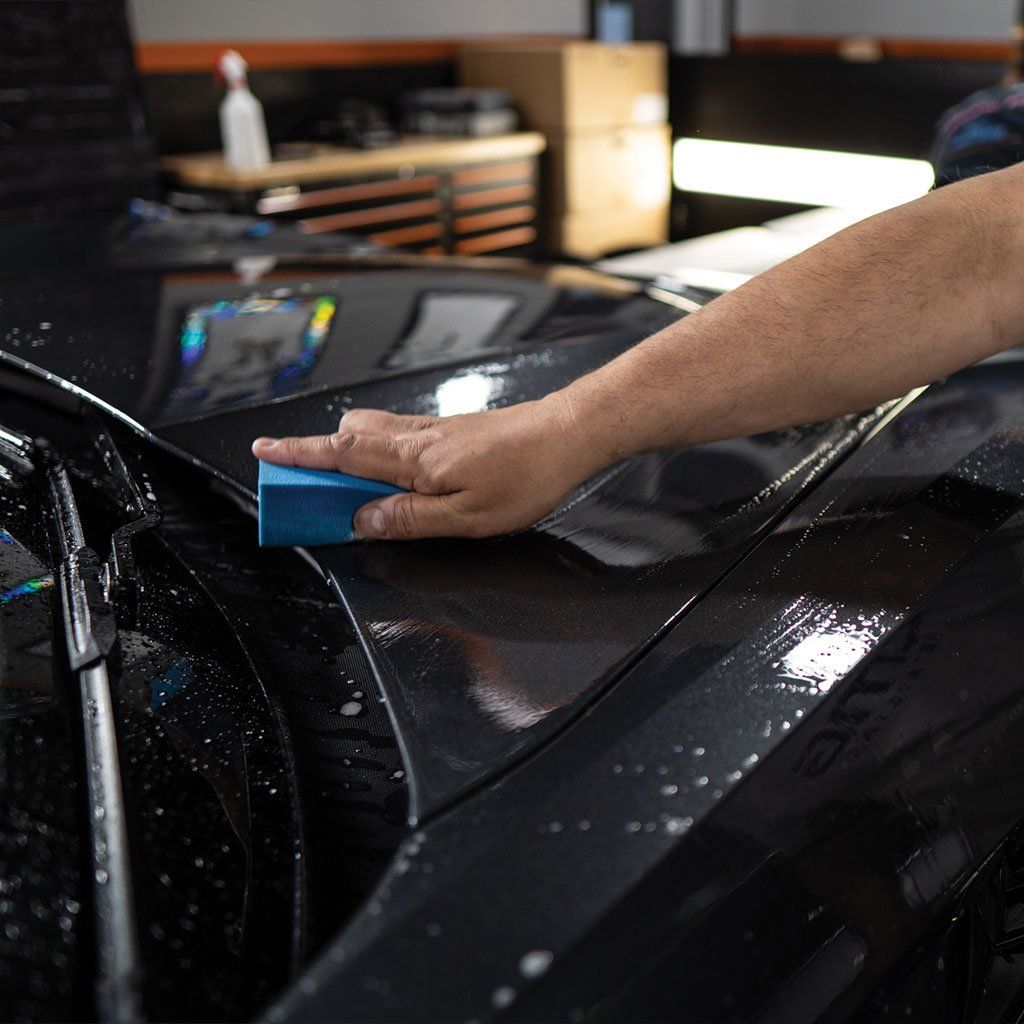 Person washing a black car with a blue sponge, suds visible.