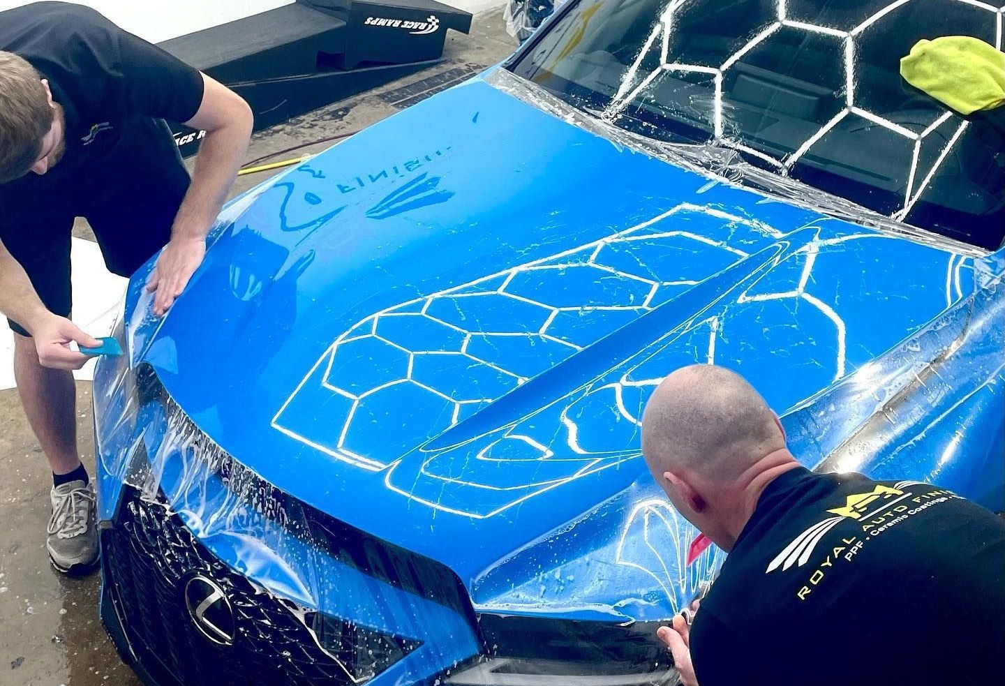 Two people applying blue protective film to a car hood, soapy water visible.