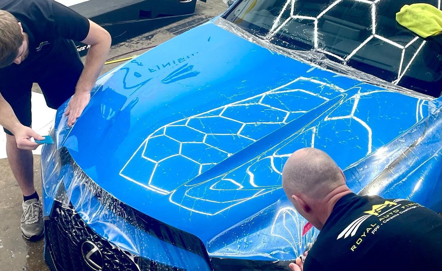 Two people applying blue protective film to a car hood. White outlines show a hexagonal pattern.
