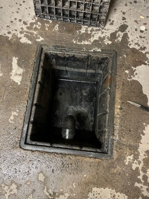 Siphon de sol noir dans une dalle de béton, caisse en plastique noir au-dessus.