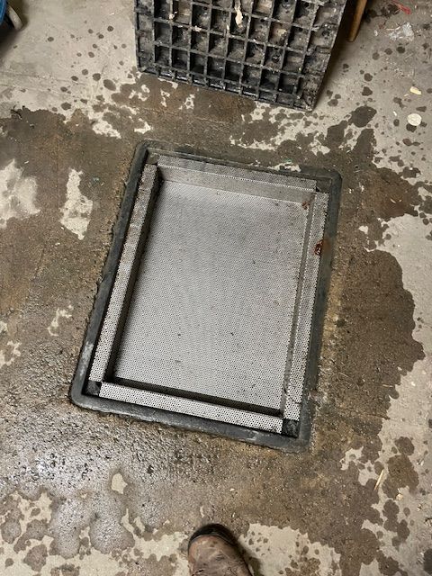 Panneau d'accès rectangulaire gris dans un sol en béton, entouré d'eau. Une caisse noire se trouve à proximité.