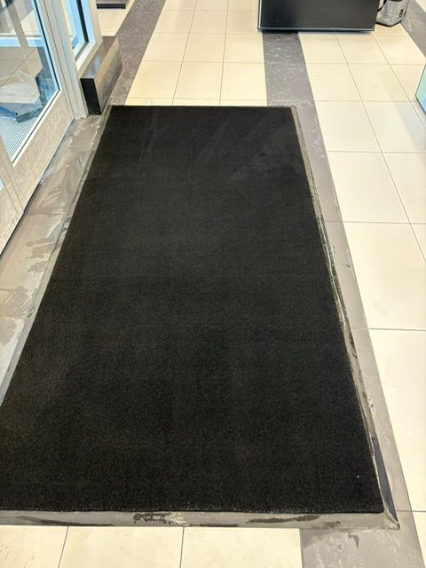 Tapis de bienvenue noir à l'intérieur d'une porte, sur un sol gris et carrelé.