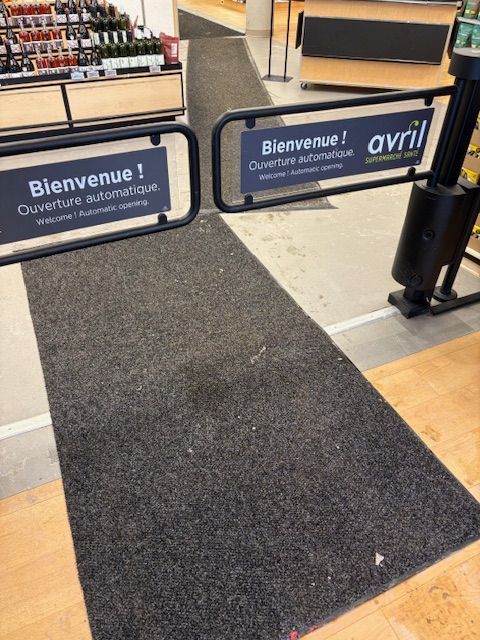 Entrée de la boutique Avril avec portes automatiques et panneaux de bienvenue. Tapis noir sur parquet.