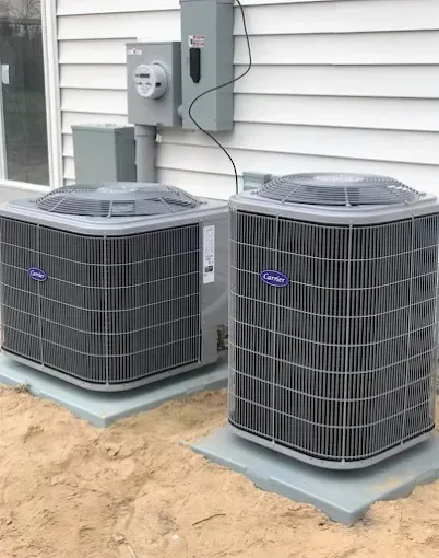 Two outdoor air conditioning units beside a house wall and electrical panel, on concrete pads.