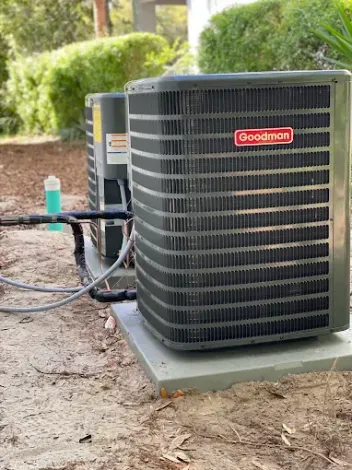 Goodman outdoor air conditioner unit on a concrete pad beside a house, with hoses attached.