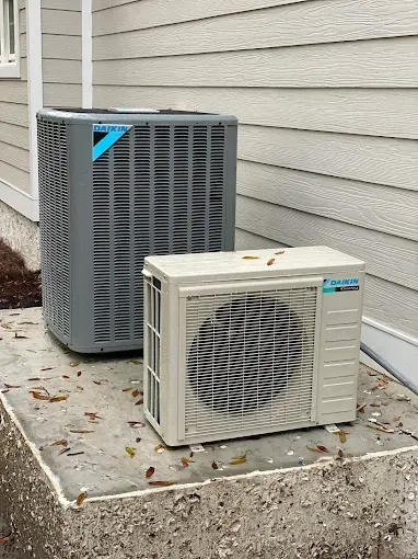Outdoor HVAC units beside a house: a large gray condenser and a small beige window AC on a concrete pad