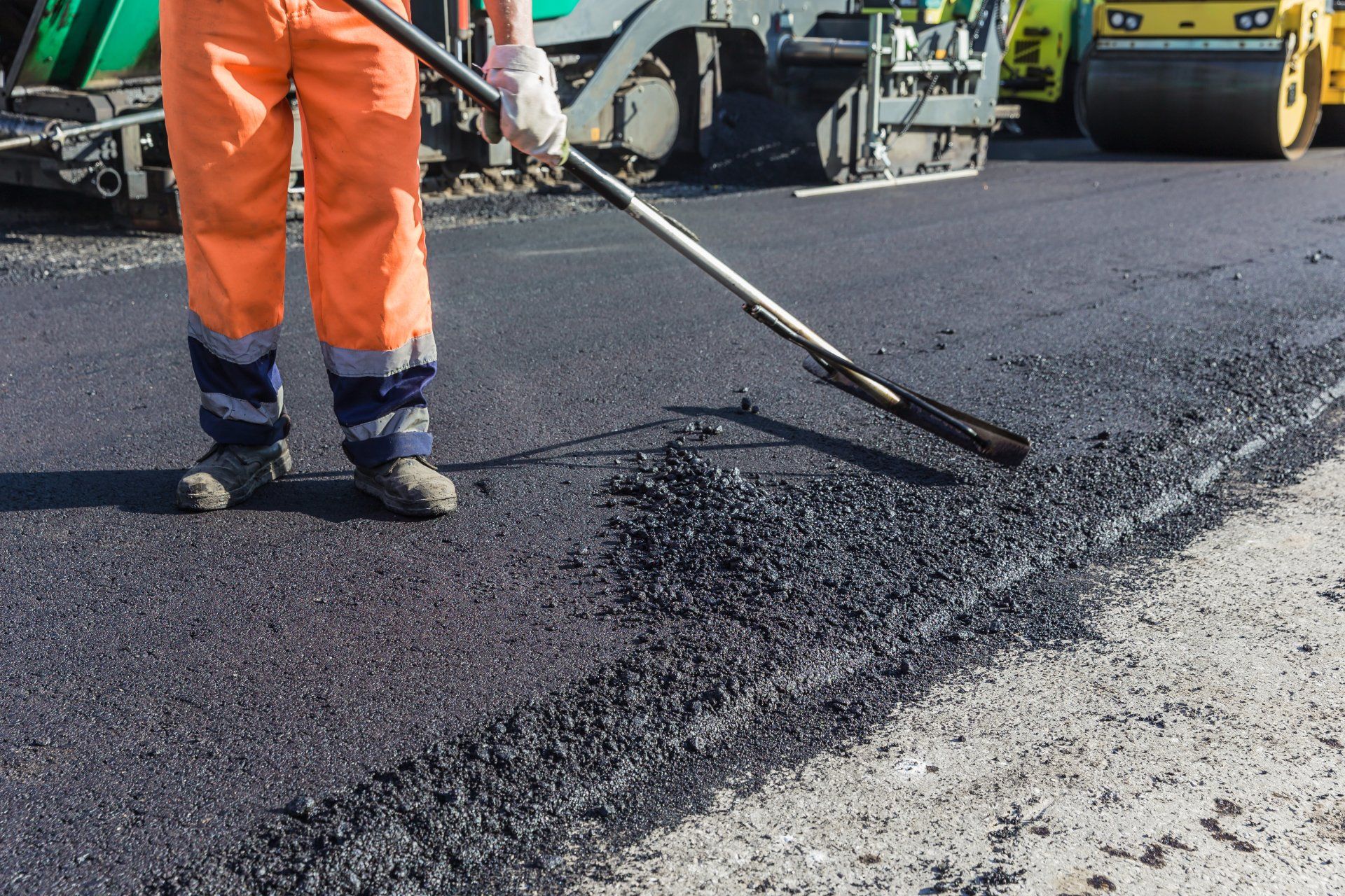 Construction worker in orange overalls smoothing asphalt with a tool, road repair.
