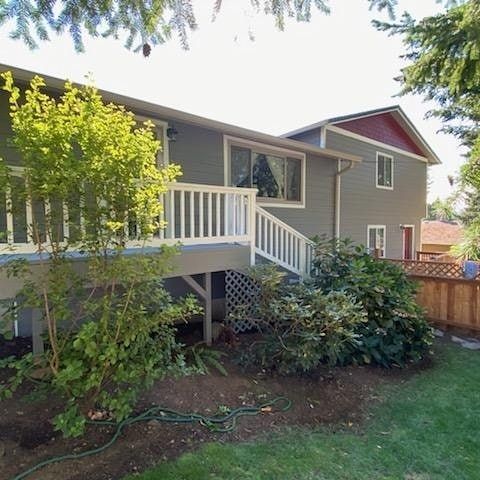 Two-story gray house with white deck and railings; lush green foliage in yard.