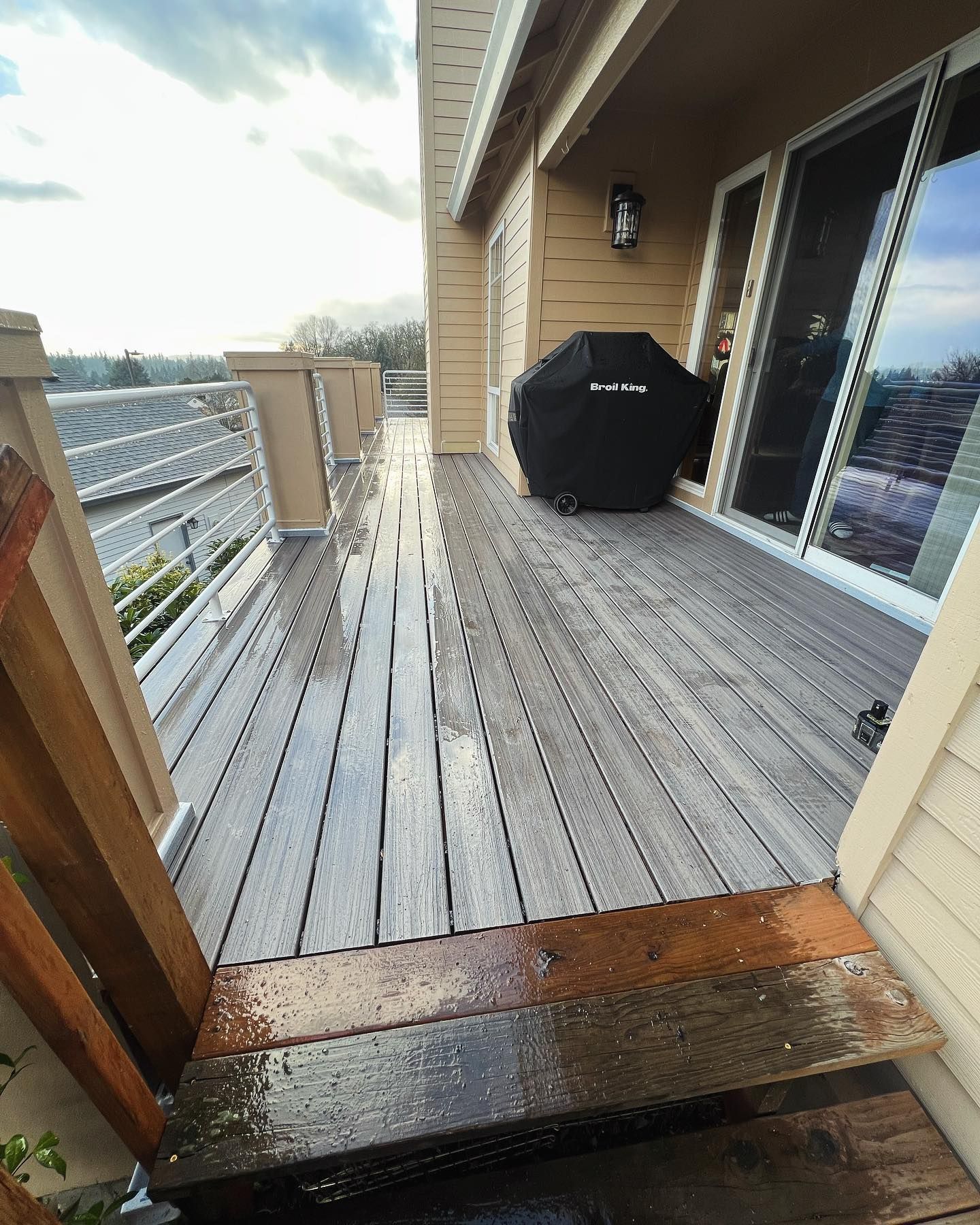 Wet wooden deck with steps, a grill, and a railing overlooking a cityscape.