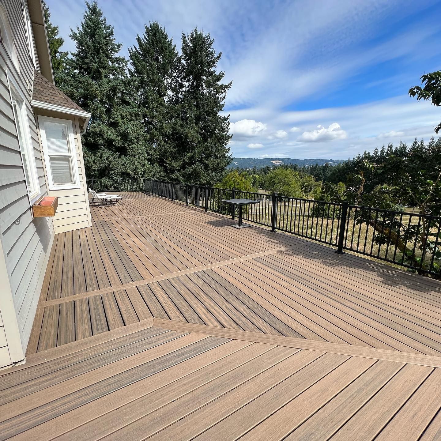 Wooden deck with black railing overlooking a green landscape on a sunny day.