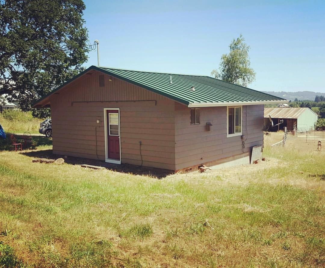 Brown house with green corrugated roof in a grassy yard under a blue sky.