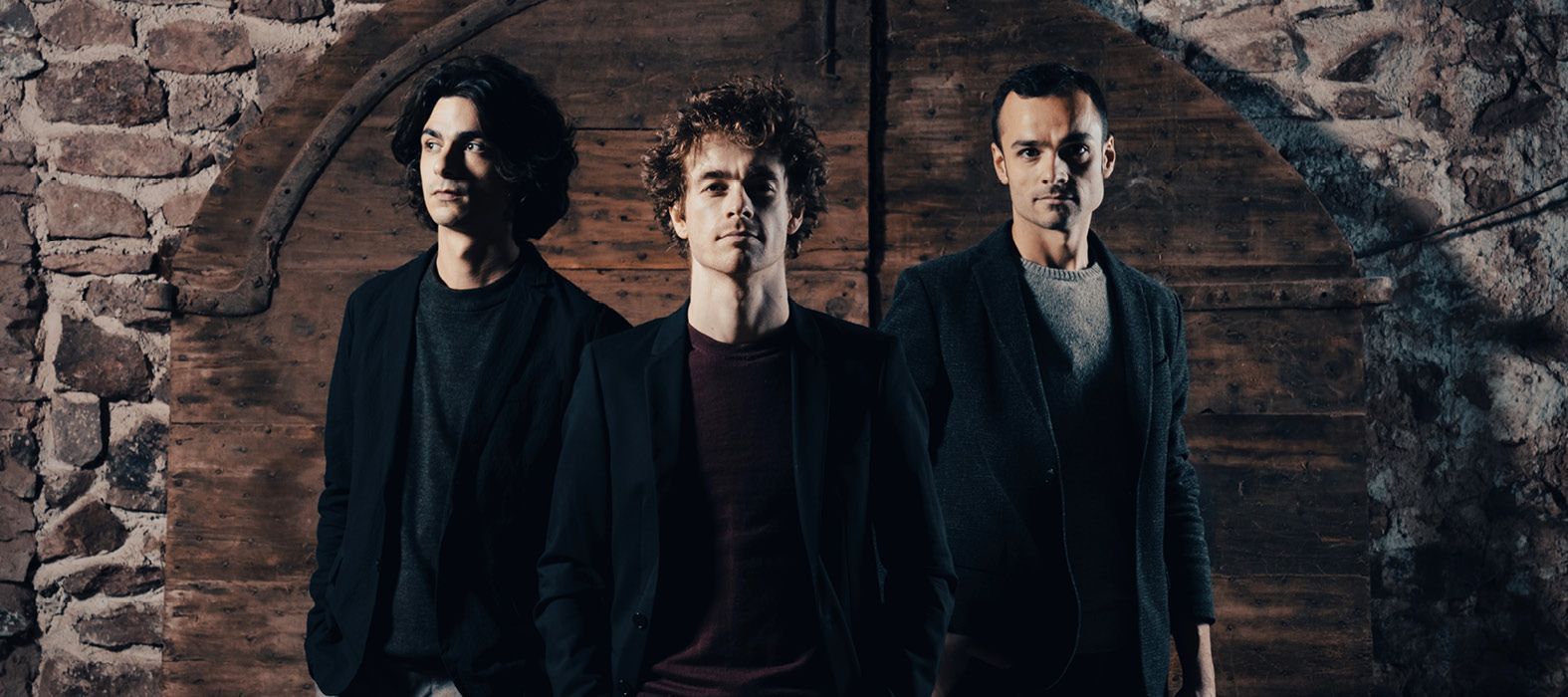 Three men in dark blazers stand in a line before a rustic stone archway, posing seriously for a portrait.