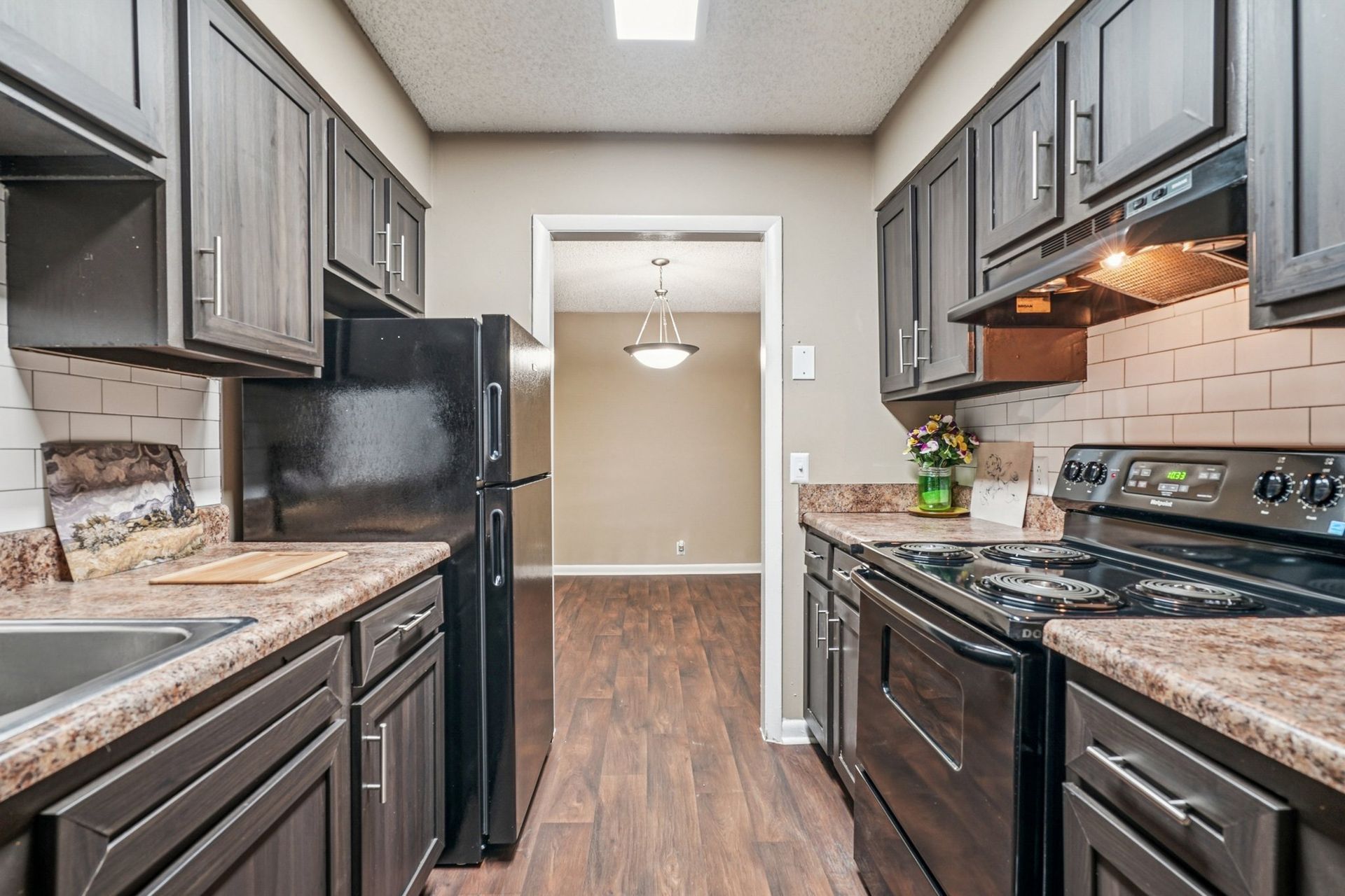 Galley kitchen with dark wood cabinets, black appliances, speckled countertops, and a doorway leading to a dining area.