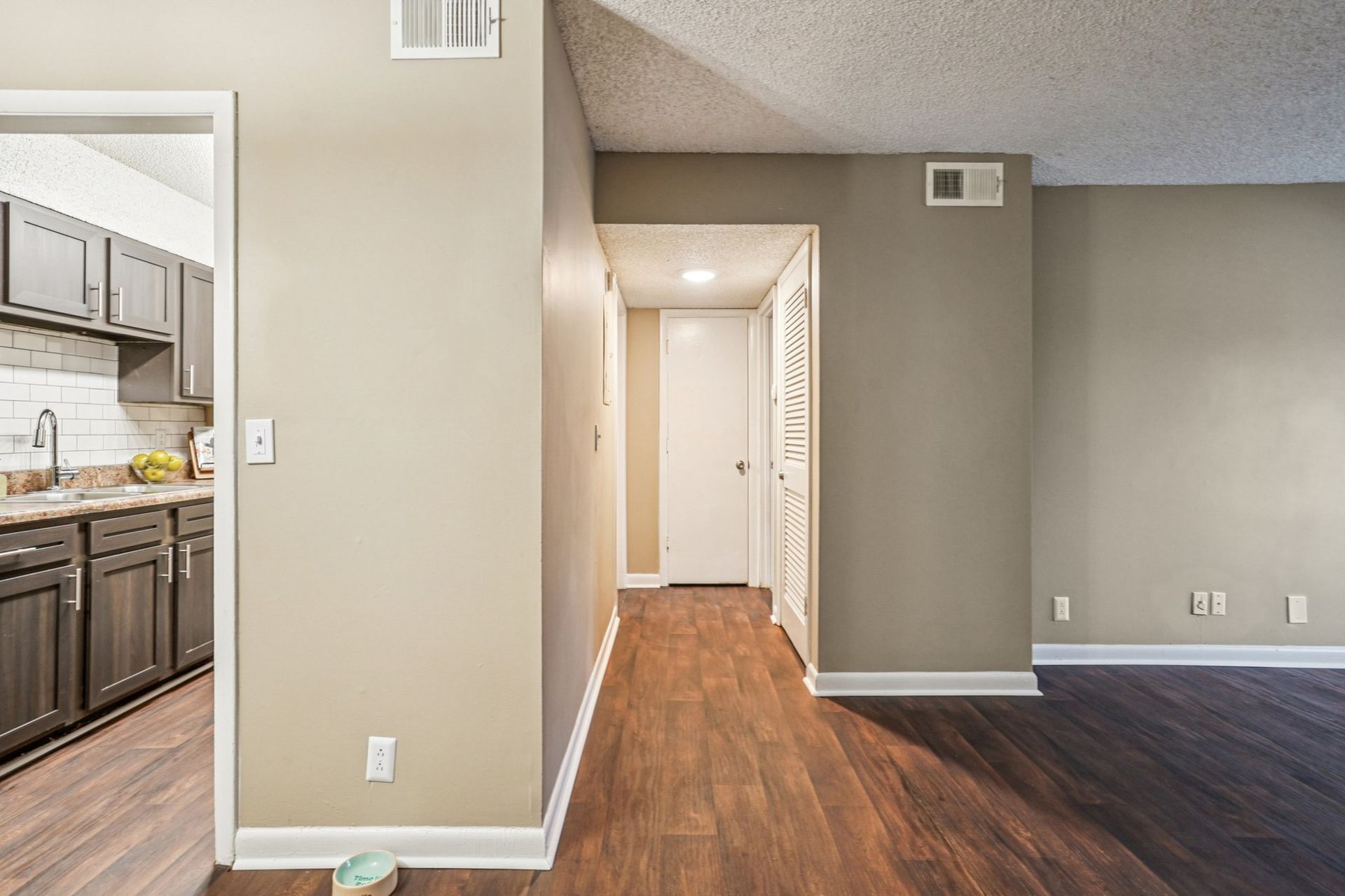 A wide-angle view of a hallway connecting a kitchen with dark wood cabinets to an adjacent living room.