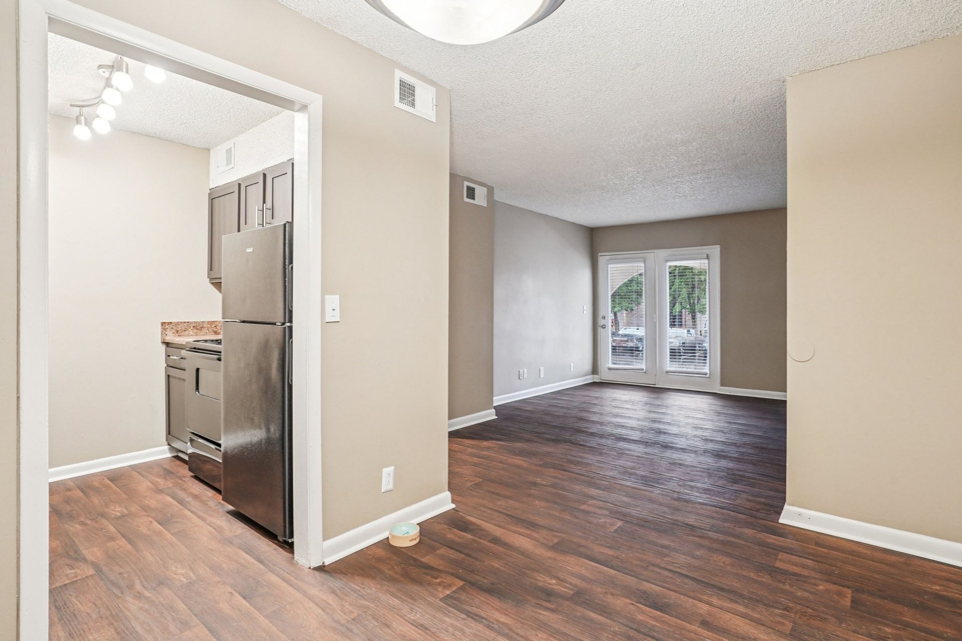 A view of an empty apartment showing a kitchen with stainless steel appliances to the left and a living area to the right.