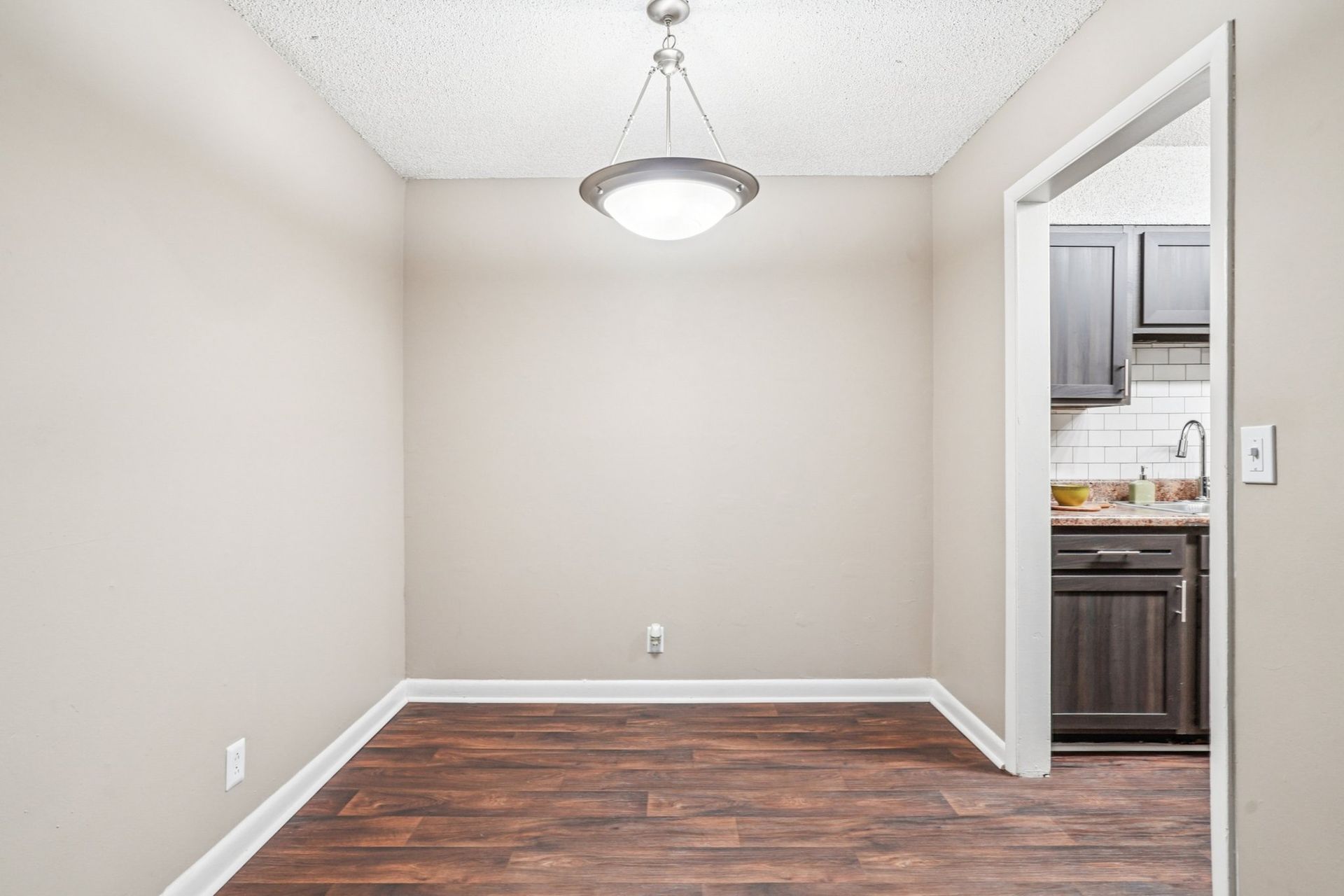 A vacant dining area with beige walls, wood-look flooring, and a modern pendant light, looking into a kitchen.