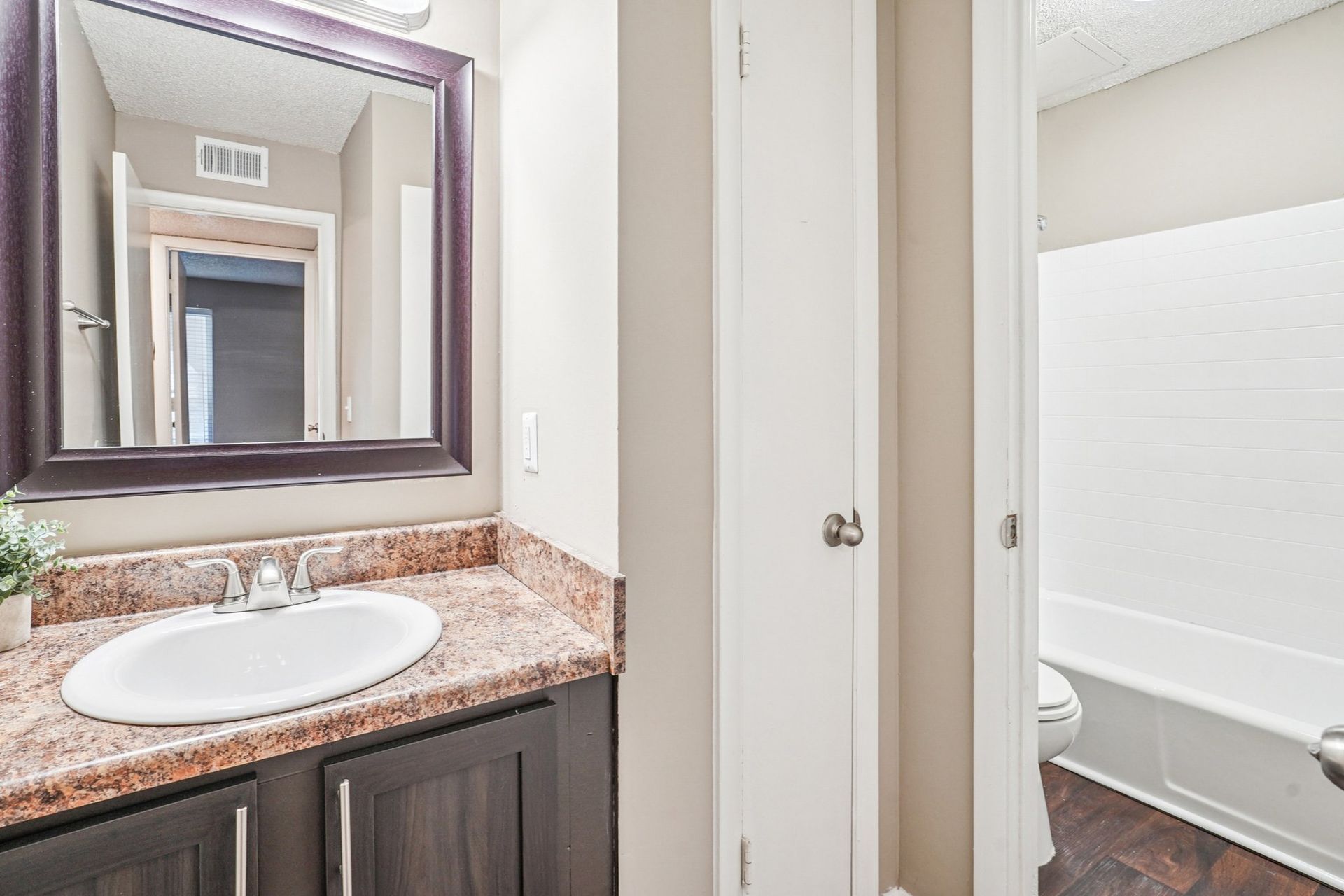 A bathroom vanity with a speckled countertop, dark cabinets, a mirror, and an adjacent door leading to a tub and toilet.