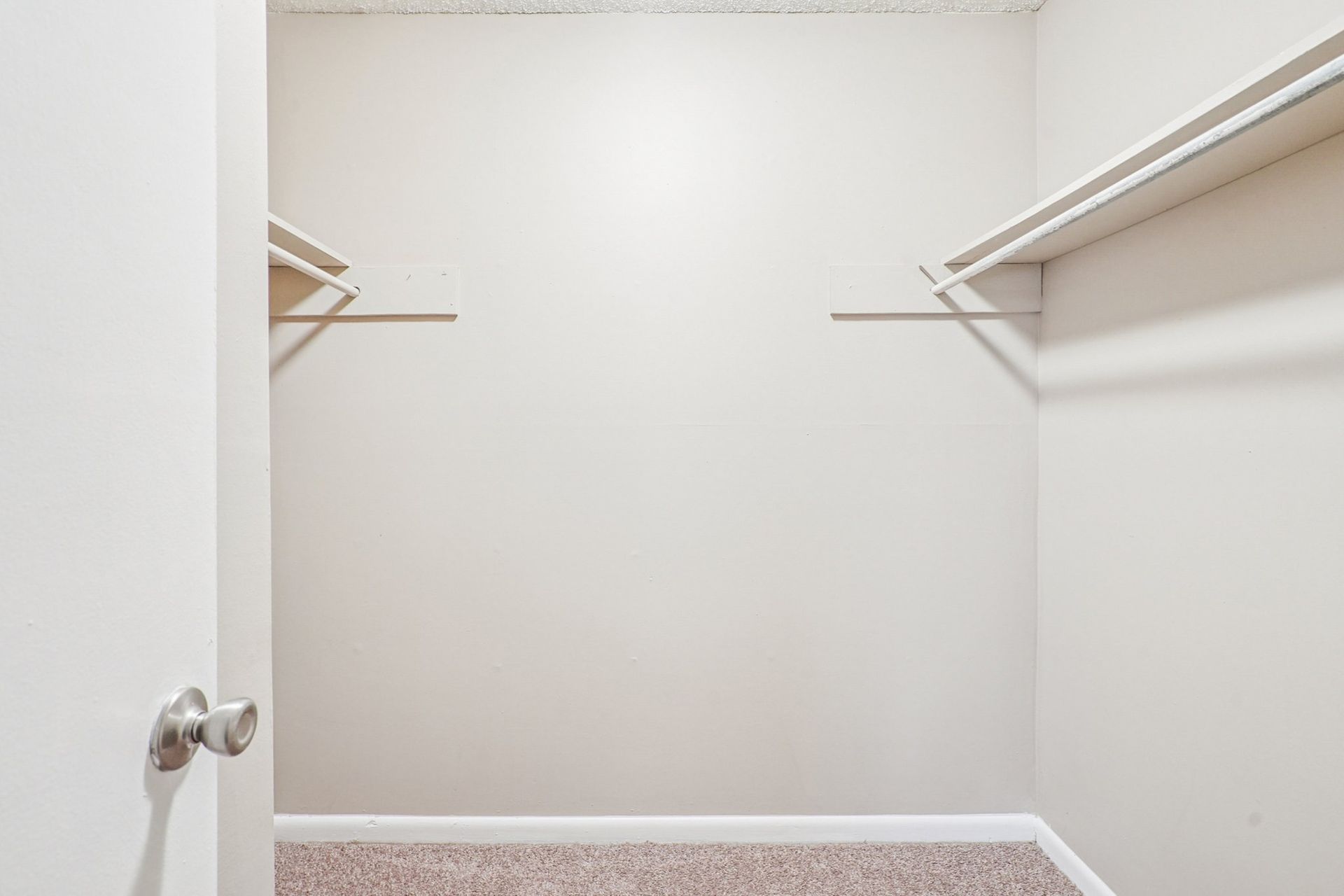 An empty walk-in closet with beige walls, tan carpeting, and white shelving brackets on either side.